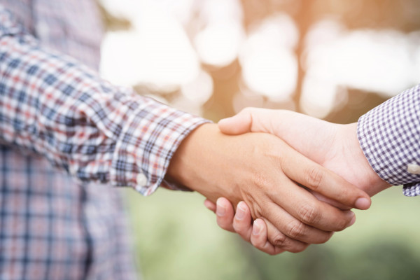 A close up of two white men wearing checked button-down shirts shake hands outdoors.